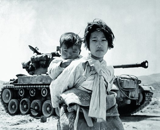 1200px-With_her_brother_on_her_back_a_war_weary_Korean_girl_tiredly_trudges_by_a_stalled_M-26_tank,_at_Haengju,_Korea_HD-SN-99-03144.png